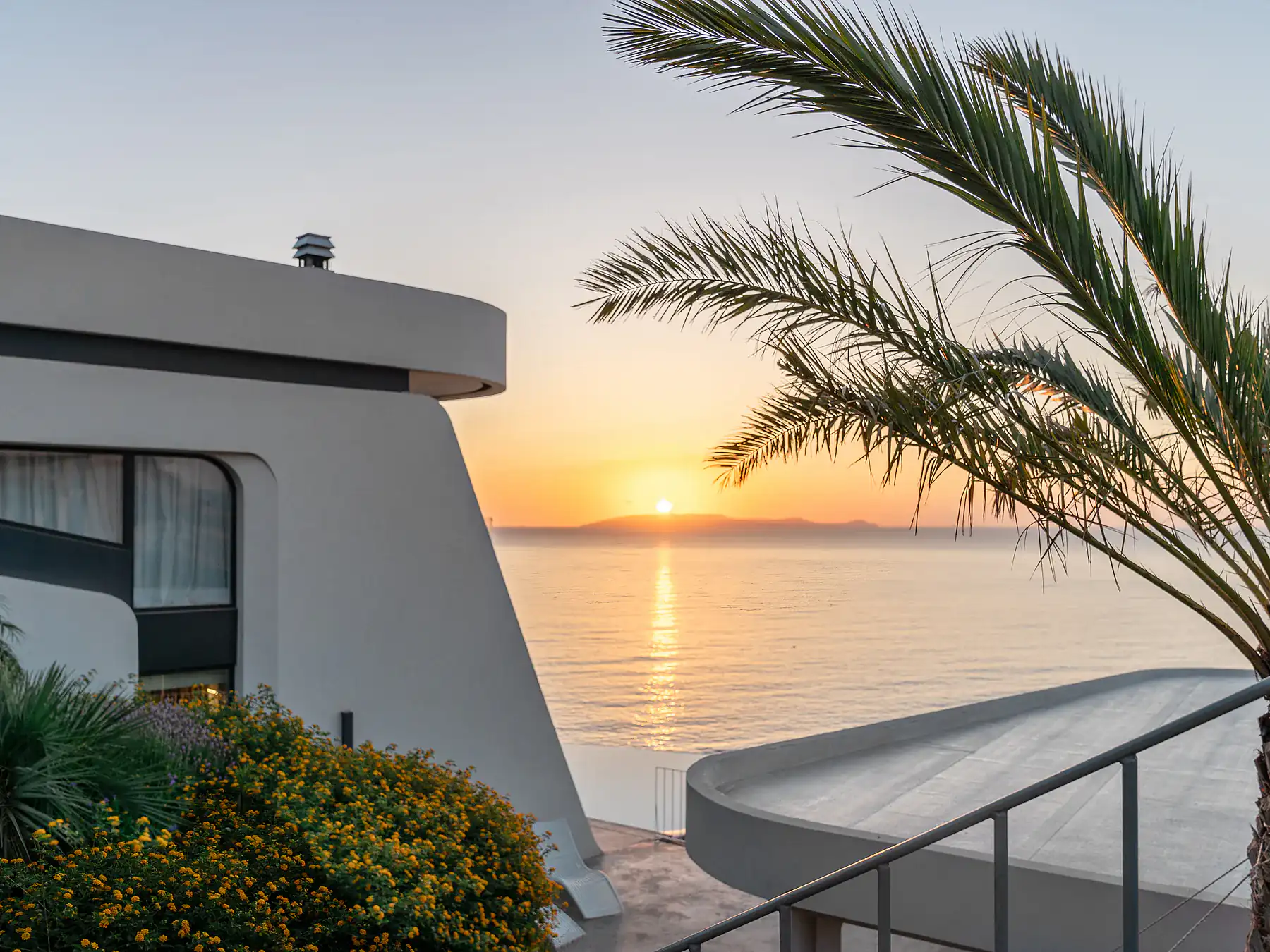 Evening view from villa terrace with palm tree and sun setting over the Aegean Sea.
