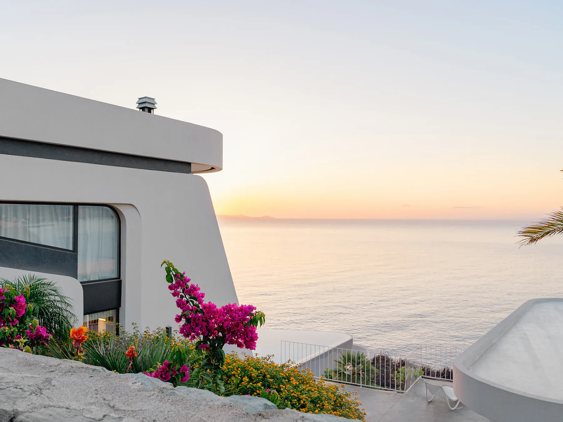 Modern villa terrace adorned with colorful flowers overlooking the calm Aegean Sea at sunset.